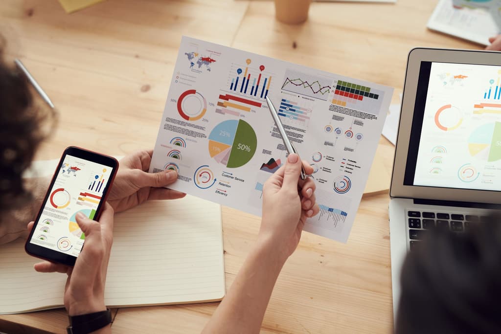 Hands holding a phone and pen over printed charts and survey sheets beside a laptop showing graphs