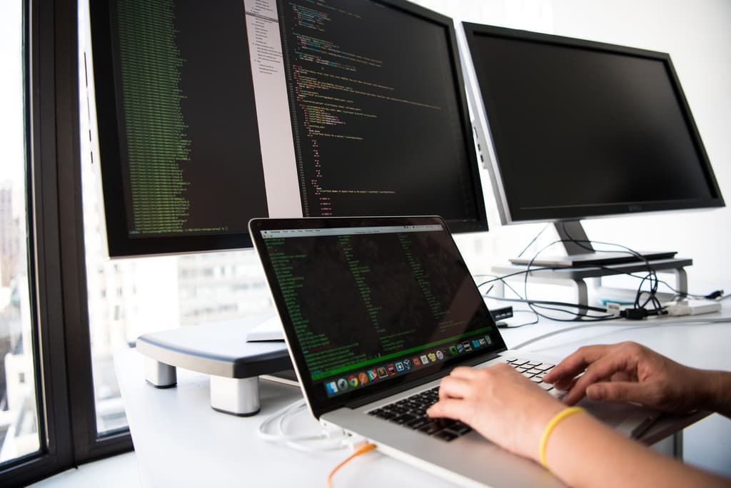 A person typing on a laptop in front of two monitors displaying code at a workstation