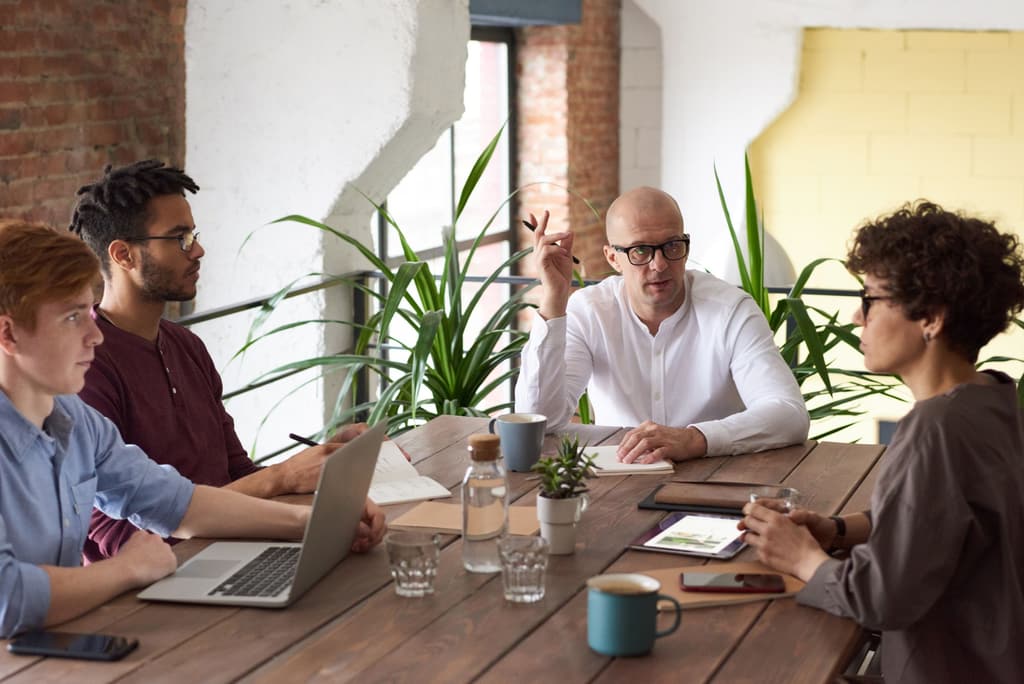 A small team in discussion around a wooden table with laptops, notebooks and plants in a bright office